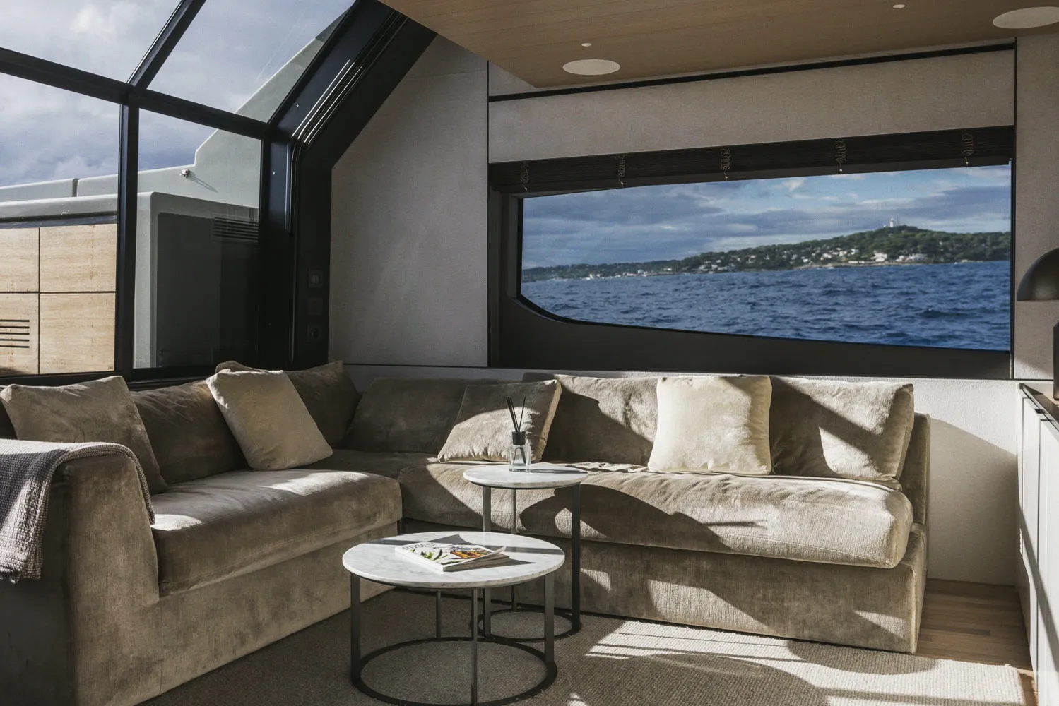 Le carré d'un yacht : banquette d'angle en velours et tables basses en marbre, une baie panoramique ouverte sur la mer et une côte boisée.