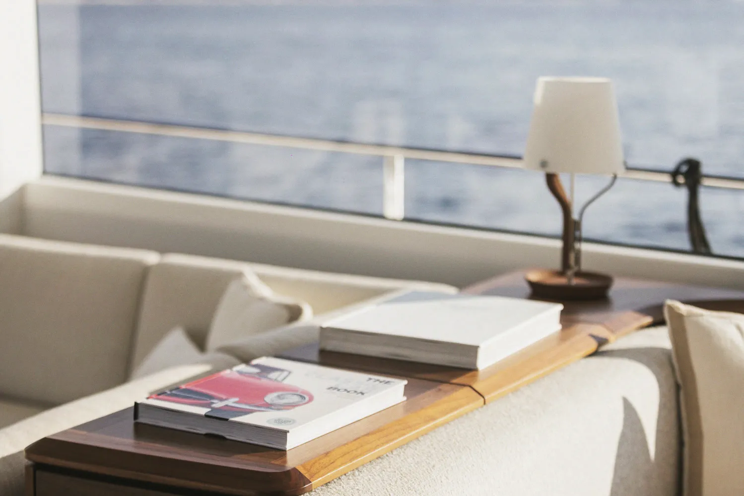 Détail du salon : lampe de table, livres et canapé clair devant la baie.