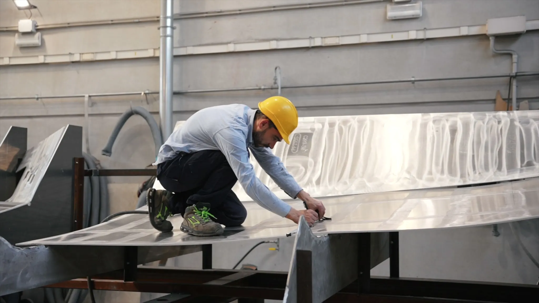 Un homme en casque de chantier, agenouillé sur une grande tôle d'aluminium posée sur des tréteaux d'acier, y trace un repère ; derrière lui, la paroi nervurée de l'atelier, ses gaines et un tuyau enroulé.