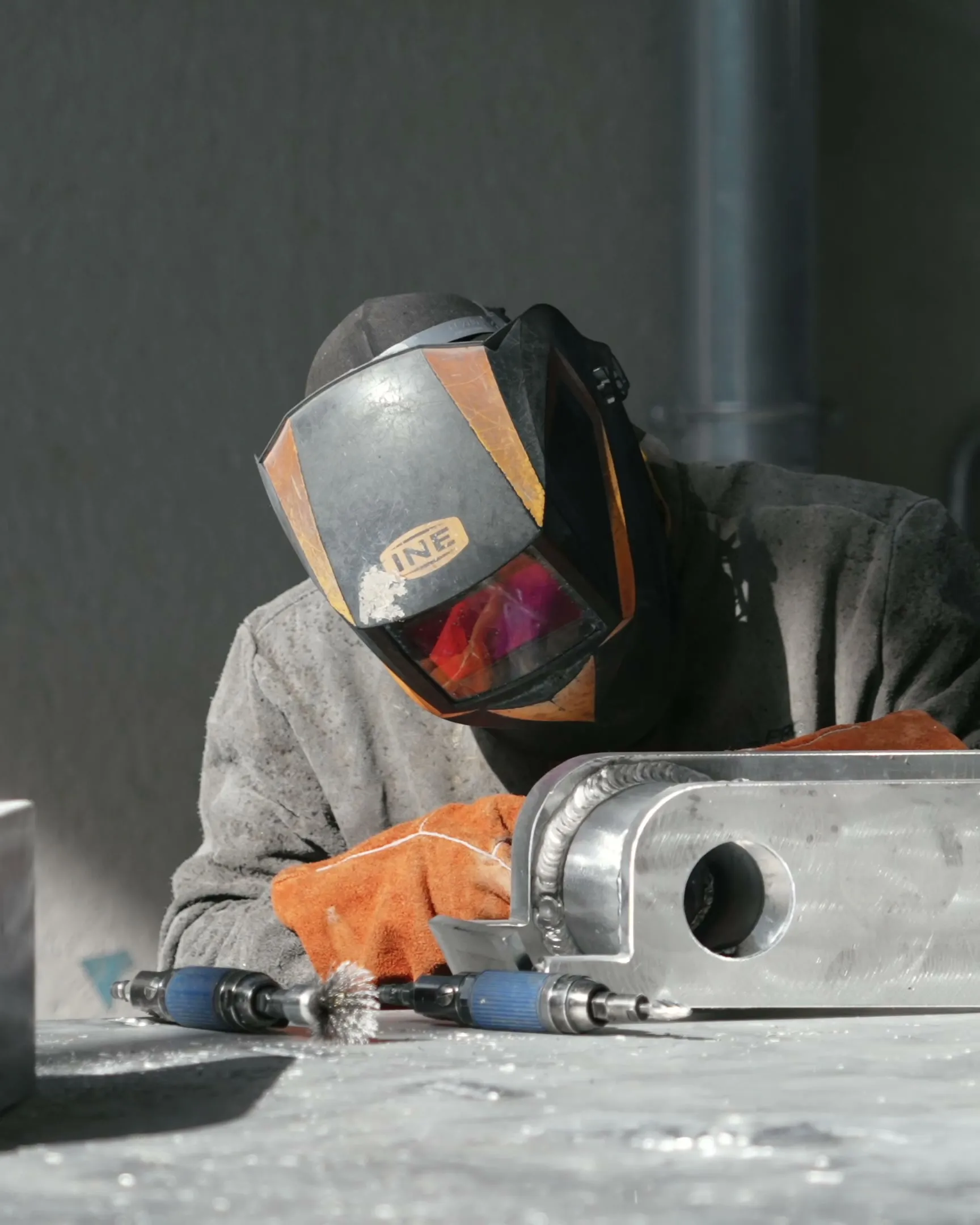 Un soudeur penché sur une pièce d'aluminium, masque baissé : le cordon de soudure court le long de l'arête, un gant de cuir posé à côté, deux outils pneumatiques sur l'établi.