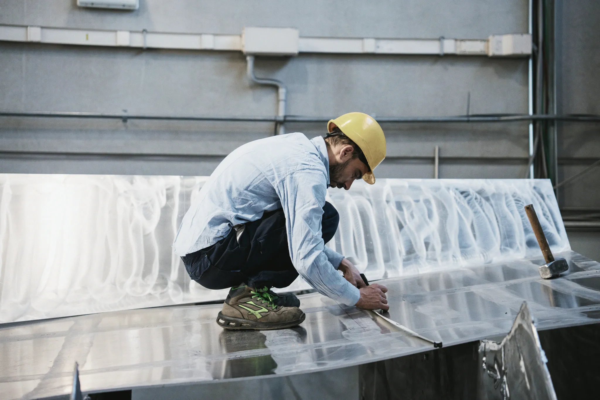 Dans un hall de chantier, un ouvrier casqué accroupi sur une tôle d'aluminium trace un repère à la règle.