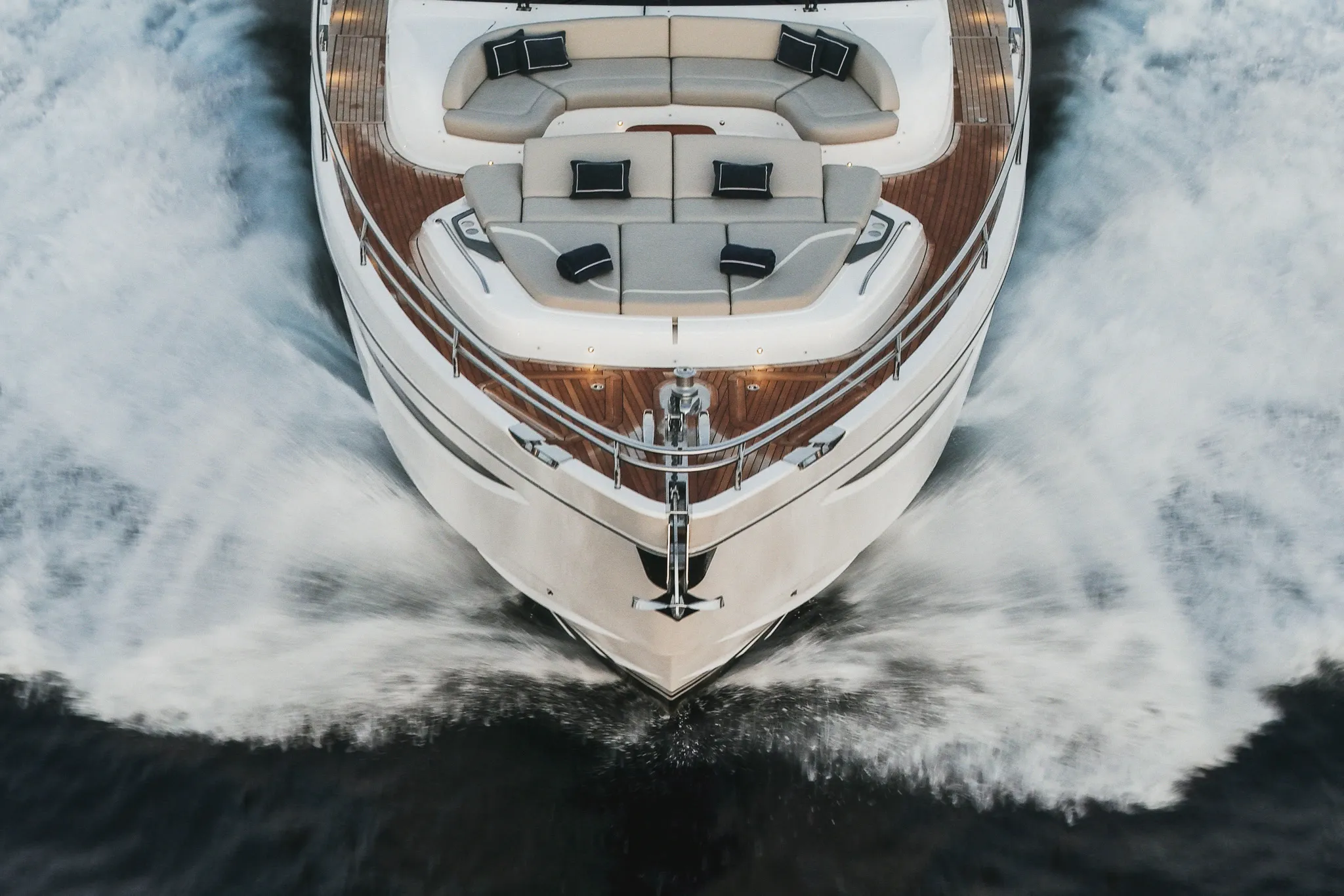 Étrave d'un yacht blanc vue de haut, en marche : plage avant en teck, bain de soleil aux coussins clairs, écume ouverte de part et d'autre de la coque.
