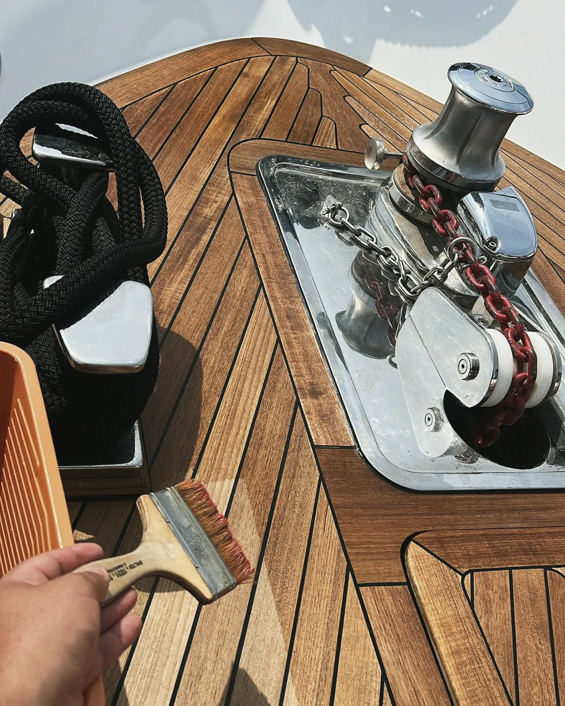 Une main passe une brosse sur le teck du pont avant : guindeau inox et chaîne d'ancre à côté, cordage lové sur son taquet, seau posé au pied.
