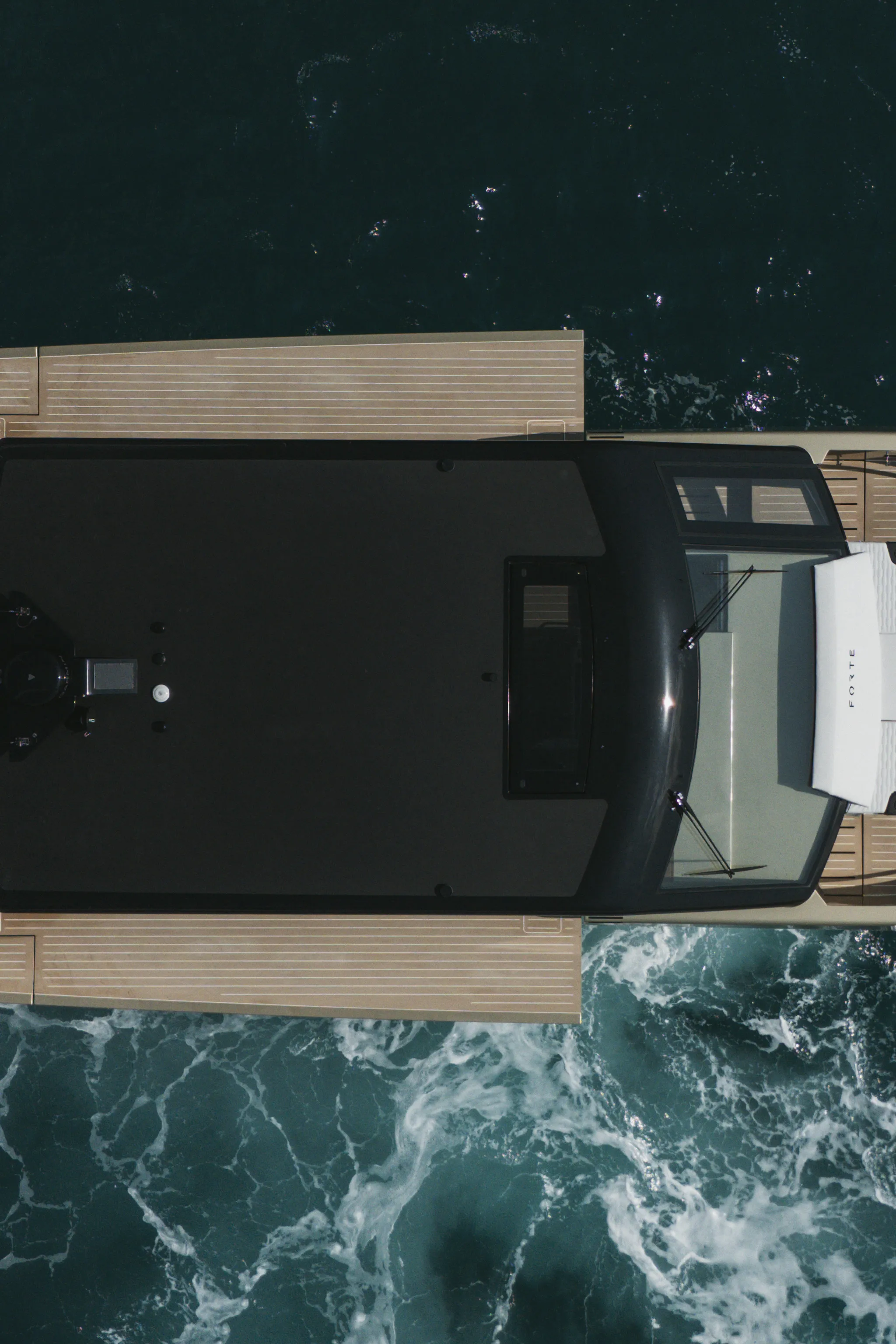 Pont d'un yacht vu à la verticale : hard-top sombre, passavants en teck de part et d'autre, écume au pied du cadre.