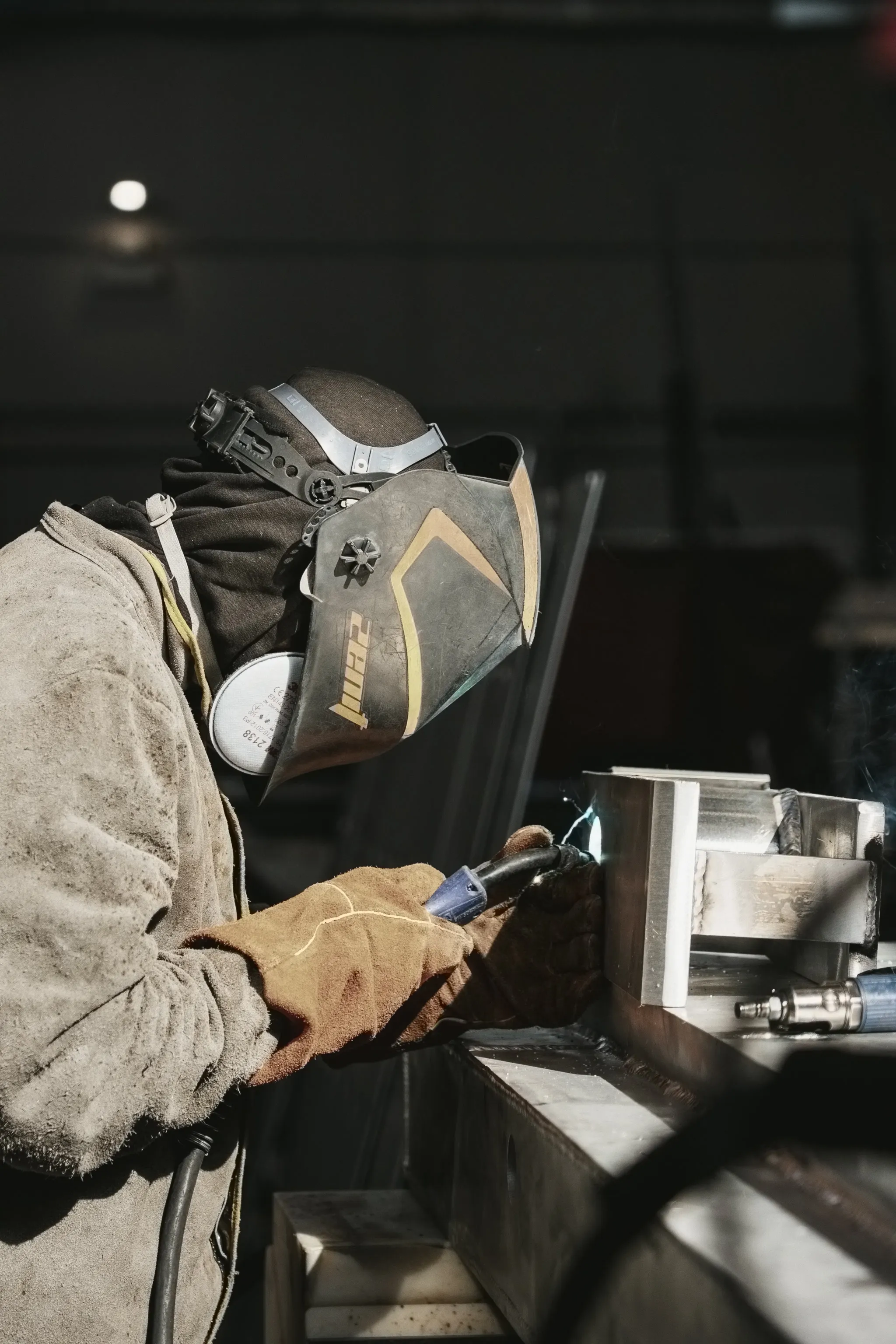 Un soudeur en veste de cuir, masque de soudage baissé, soude à la torche un assemblage d'aluminium posé sur l'établi : l'arc est amorcé, halo bleu et projections au point de soudure.