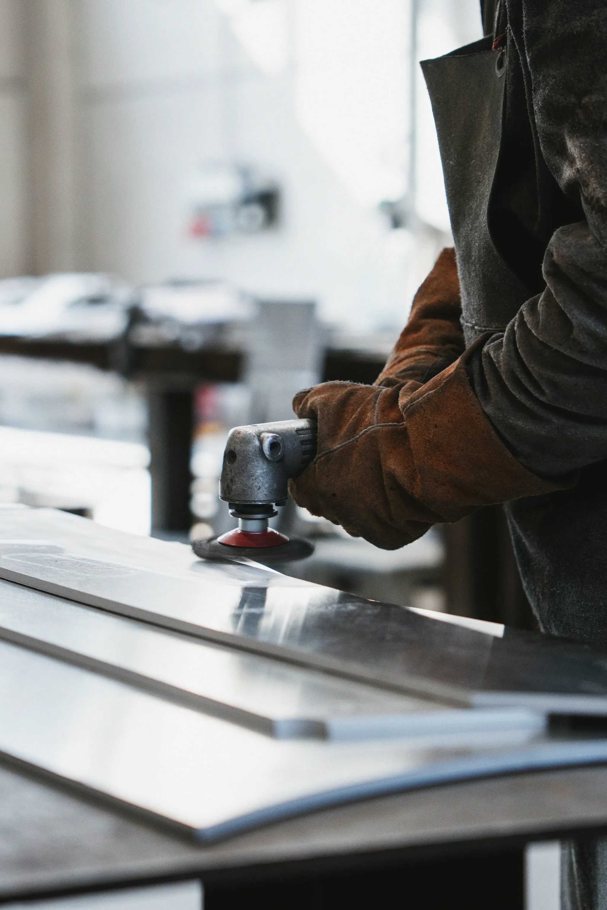 Une main gantée guide une ponceuse à disque sur une tôle d'aluminium, l'atelier flou derrière.
