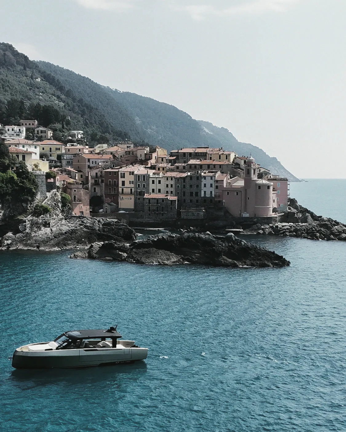 Yacht à moteur au mouillage sur une eau turquoise, devant un village bâti sur un promontoire rocheux : façades serrées, toits de tuile, colline boisée en arrière-plan.