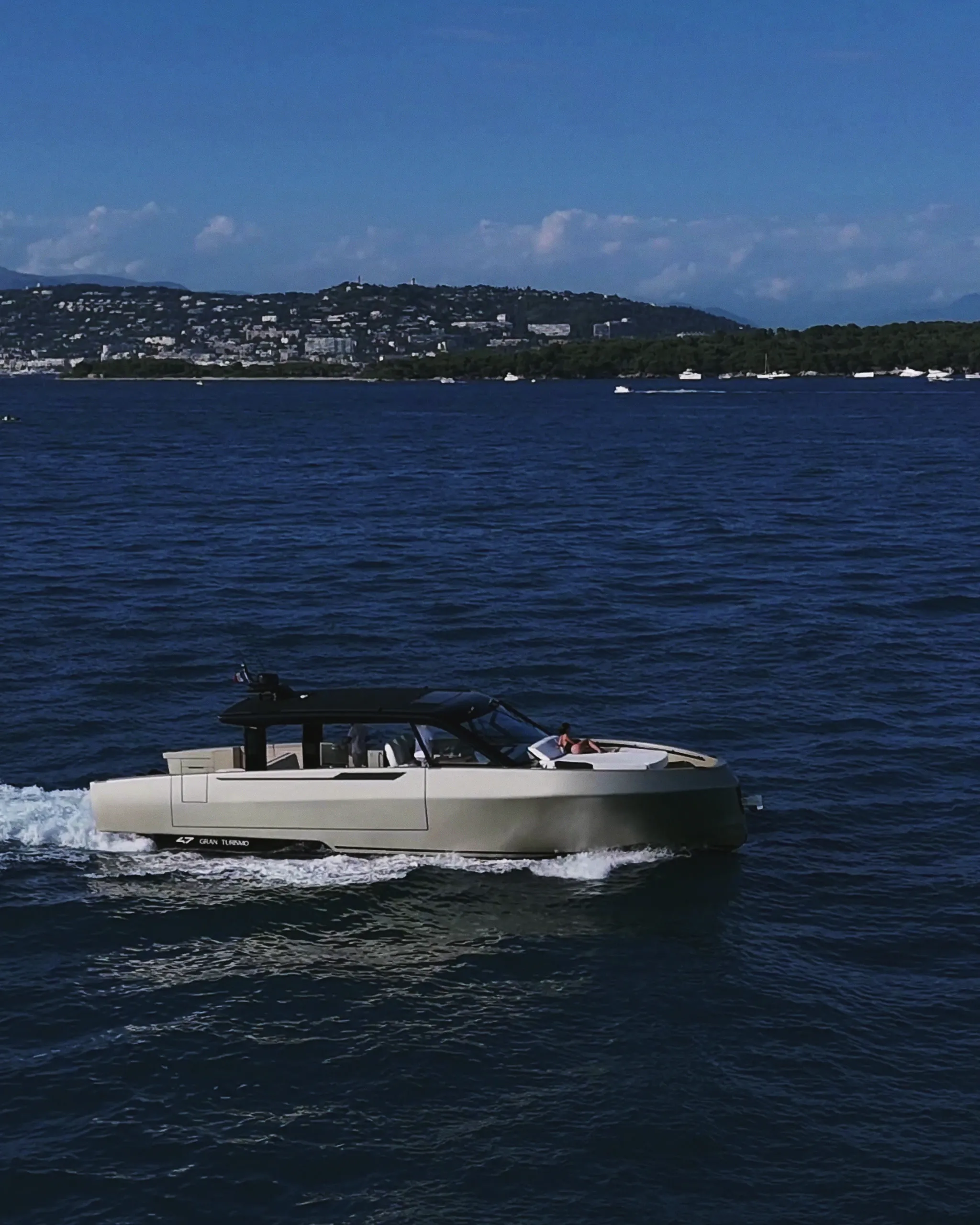Yacht à moteur en marche sur une mer ouverte, coque claire et hard-top sombre ; au loin, une côte boisée, des collines et quelques bateaux au mouillage.