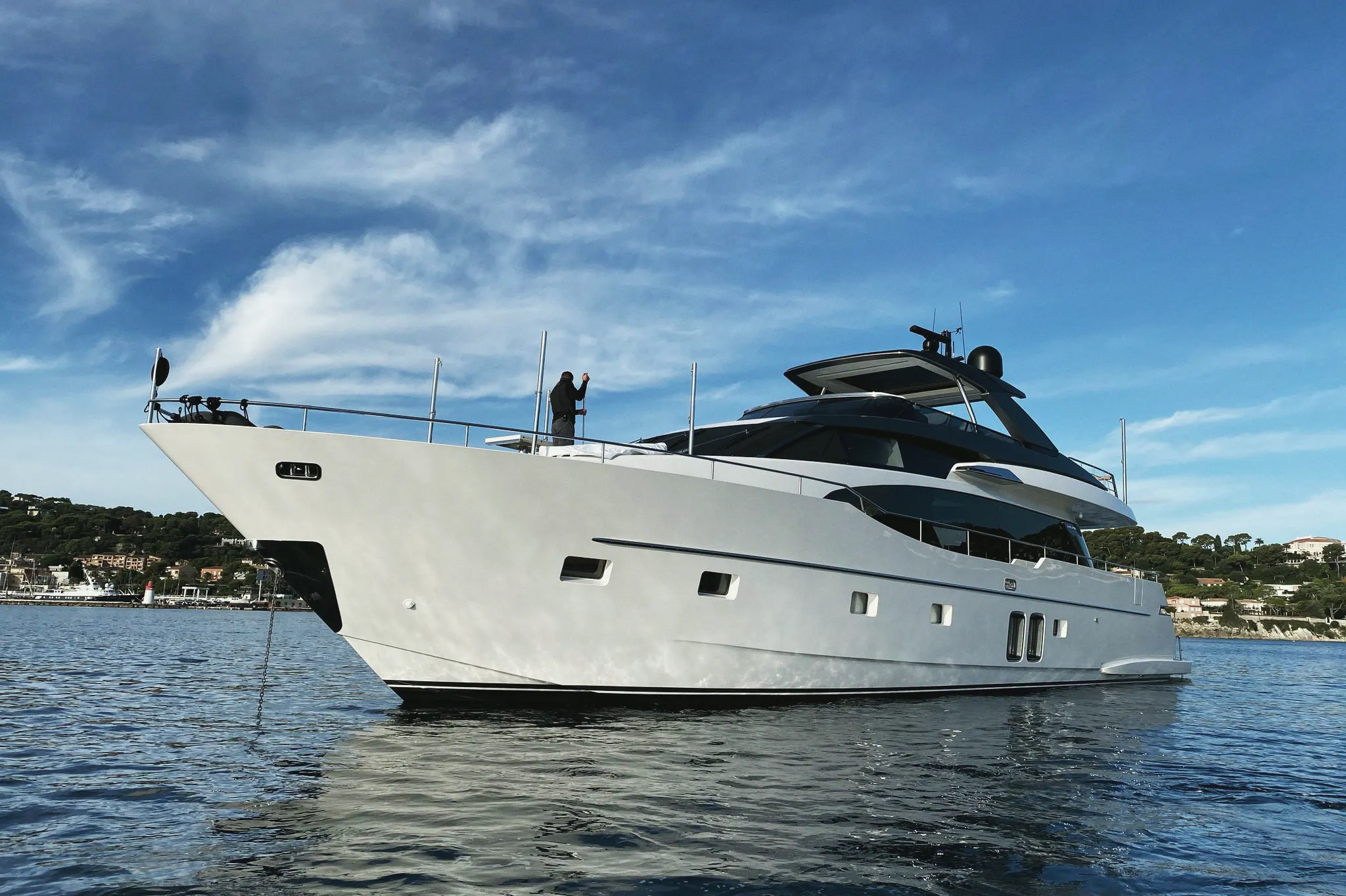Un yacht au mouillage vu de trois quarts avant, la chaîne d'ancre plongée dans une mer calme, une personne sur la plage avant, la côte en arrière-plan.