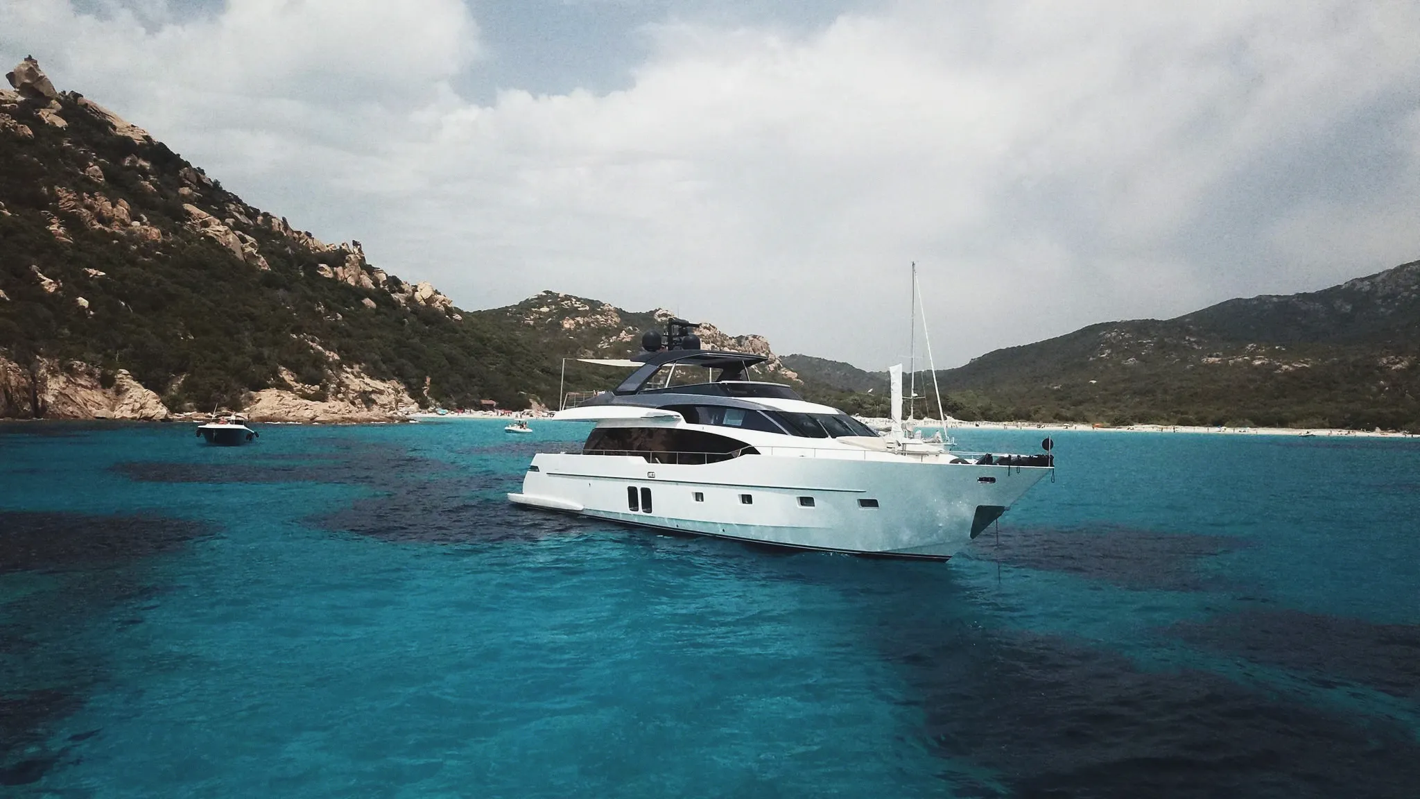 Un yacht au mouillage dans une baie aux eaux turquoise, entre collines rocheuses et plage.
