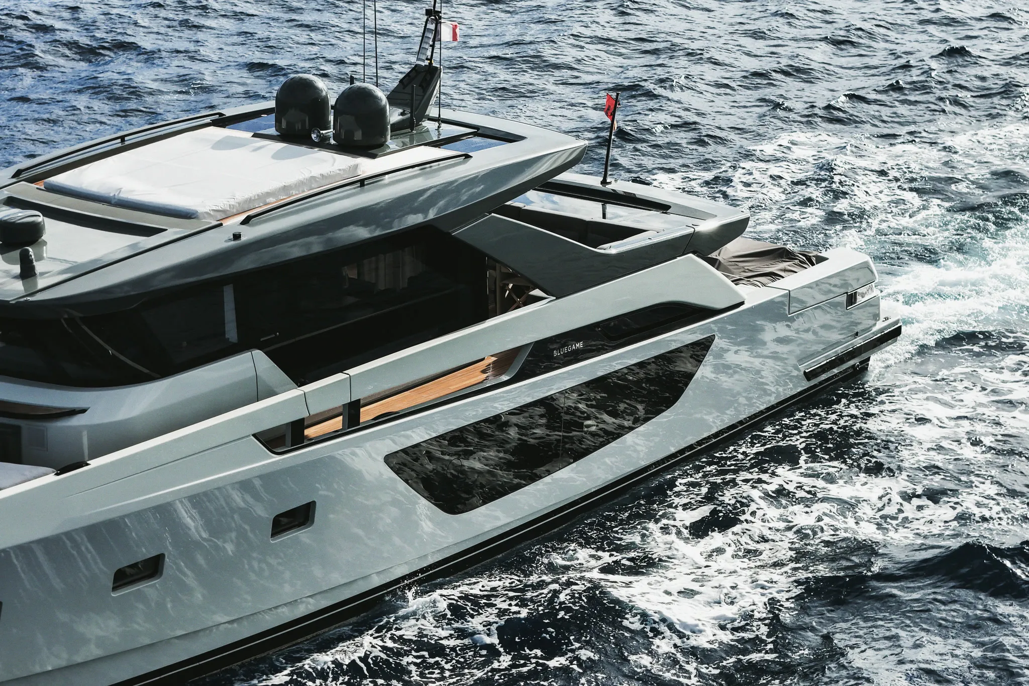 Yacht à moteur vu de haut, en marche : pont supérieur clair et dômes de communication, baies vitrées sombres, sillage d'écume le long de la coque.